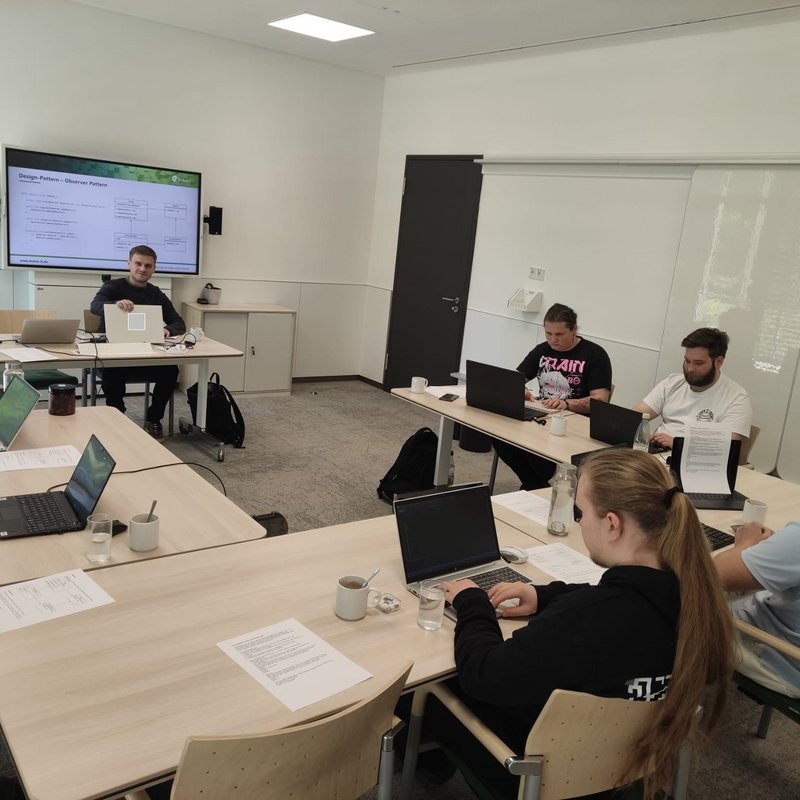 Auszubildende der kubus IT sitzen mit Laptops in einem Seminarraum und nehmen an einem IT-Lehrgang zur objektorientierten Programmierung teil. Das Bild zeigt eine Gruppe von Auszubildenden der kubus IT in einem modernen Seminarraum. Sie sitzen mit Laptops an Tischen und arbeiten konzentriert während eines Lehrgangs zur objektorientierten Programmierung. Im Hintergrund ist ein Dozent zu sehen, der Inhalte auf einem großen Bildschirm präsentiert. Die Atmosphäre wirkt fokussiert und praxisnah.