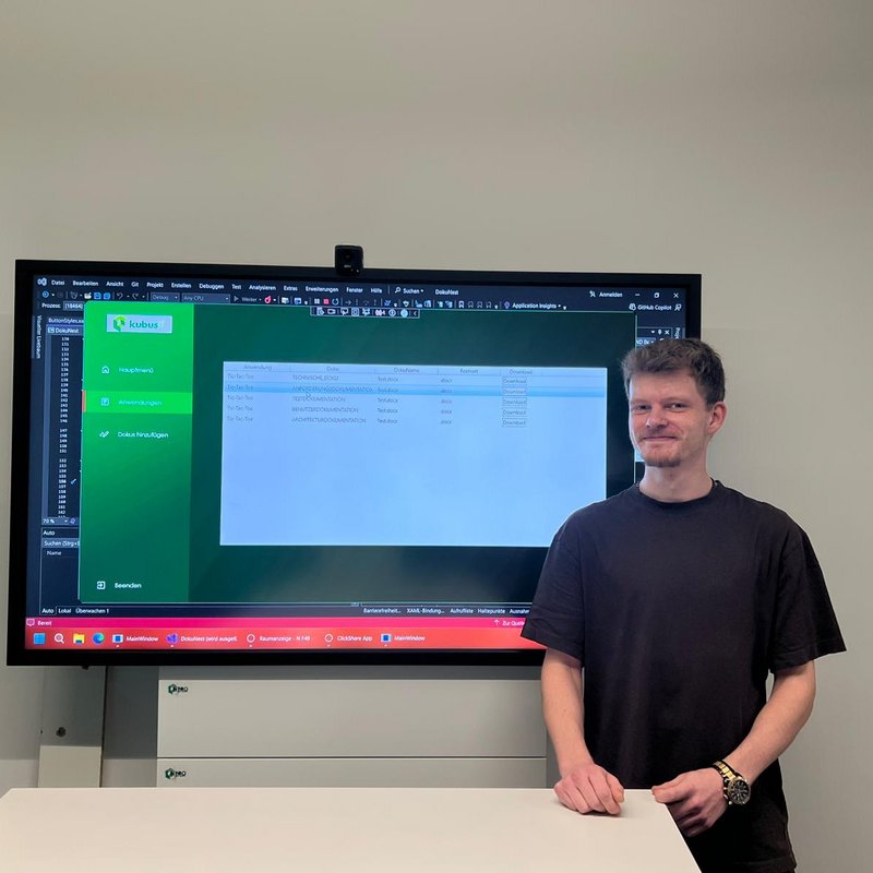 Vorstellung des „Doku-Checker“-Azubiprojekts im Unternehmen Der Auszubildende Philipp steht vor einem großen Bildschirm und präsentiert eine selbst entwickelte IT-Anwendung für Dokumentationsmanagement.