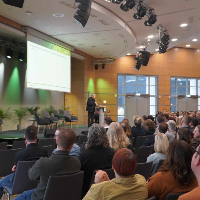 Geschäftsführer der kubus IT bei der Eröffnung der Werkstattgespräche 2026 im CongressCenter Erfurt. Eine weitwinklige Aufnahme aus dem Zuschauerraum im CongressCenter Erfurt. Im Fokus steht der Geschäftsführer der kubus IT, der auf einer Bühne an einem Rednerpult spricht. Er blickt in das Publikum, das in zahlreichen Stuhlreihen vor ihm sitzt und ihm den Rücken zuwendet. Auf der Bühne befindet sich links eine große Leinwand mit einer Präsentation sowie eine Sitzecke mit vier modernen grauen Sesseln und Grünpflanzen. Der Saal ist hell beleuchtet und verfügt über eine markante Holzvertäfelung an den Wänden.