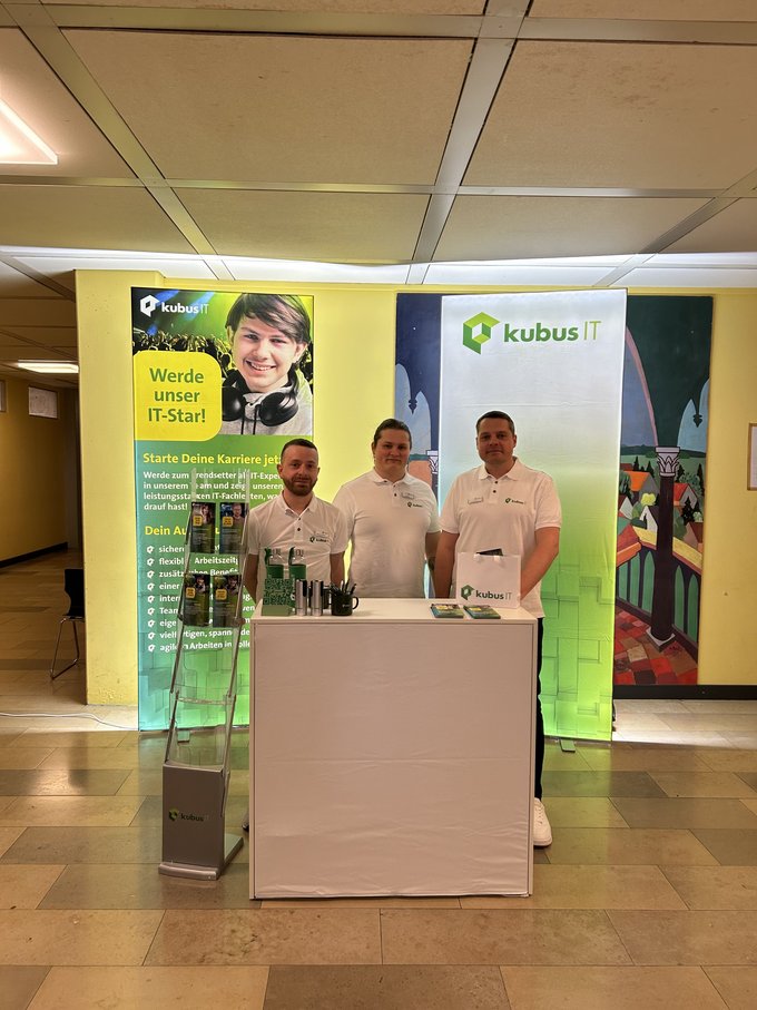 Drei Mitarbeitende von kubus IT am Messestand auf der Ausbildungsmesse in Hollfeld. Im Hintergrund Banner mit Informationen zur Ausbildung, im Vordergrund ein weißer Messetresen mit Flyern und Giveaways. kubus IT auf der Ausbildungsmesse – Team am Messestand in Hollfeld| kubusIT - Arbeitsgemeinschaft von Körperschaften des öffentlichen Rechts eGbR