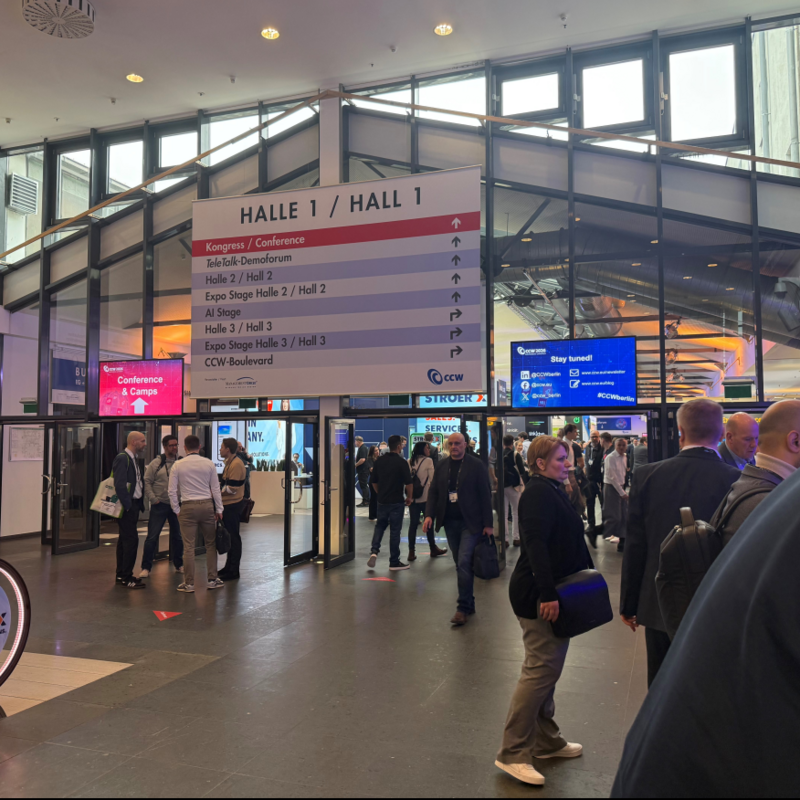 Eingangsbereich der CCW 2026 in Berlin mit Besucherandrang Im Eingangsbereich einer Messehalle stehen und gehen zahlreiche Besucherinnen und Besucher durch mehrere Glastüren. Über dem Eingang hängt ein großes Schild mit der Aufschrift „Halle 1 / Hall 1“ sowie Wegweisern zu Konferenz- und Ausstellungsbereichen. Digitale Bildschirme und Messestände sind im Hintergrund sichtbar, während sich Menschen in Business-Kleidung unterhalten und orientieren.