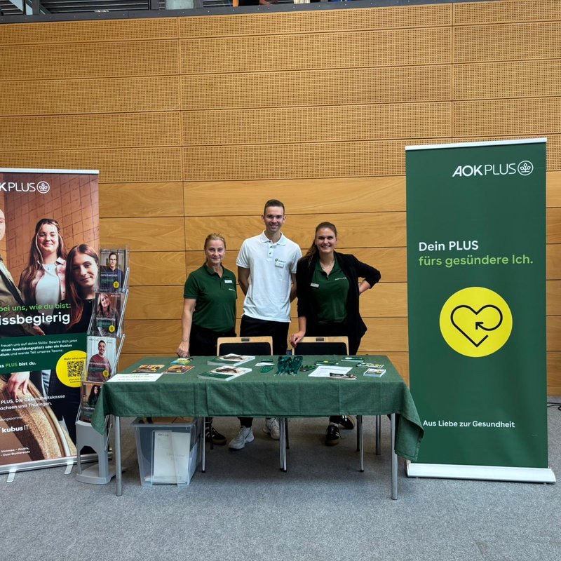 Messestand von AOK PLUS und kubus IT auf der Jobchance Messe Altenburg Drei Personen stehen hinter einem Messestand mit grünem Tischdecken, Flyern und Werbematerialien. Links und rechts sind große Roll-Ups von AOK PLUS mit den Botschaften „Sei bei uns, wie du bist: wissbegierig“ und „Dein PLUS fürs gesündere Ich“. Im Hintergrund ist eine moderne Messehalle mit Holzverkleidung zu sehen.