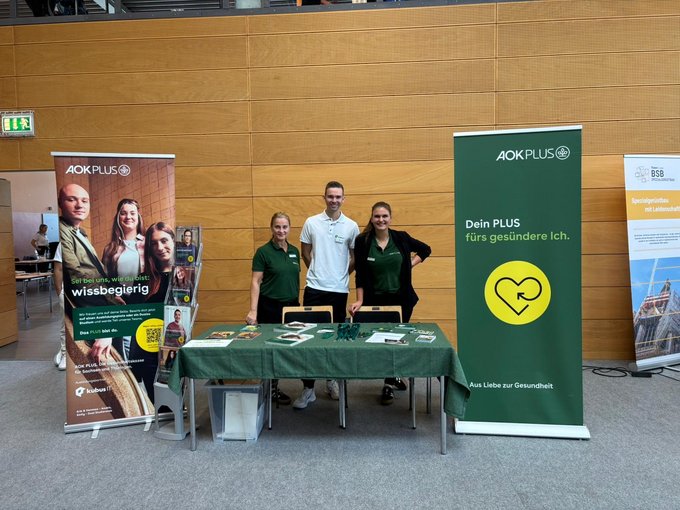 Messestand von AOK PLUS und kubus IT auf der Jobchance Messe Altenburg Drei Personen stehen hinter einem Messestand mit grünem Tischdecken, Flyern und Werbematerialien. Links und rechts sind große Roll-Ups von AOK PLUS mit den Botschaften „Sei bei uns, wie du bist: wissbegierig“ und „Dein PLUS fürs gesündere Ich“. Im Hintergrund ist eine moderne Messehalle mit Holzverkleidung zu sehen.