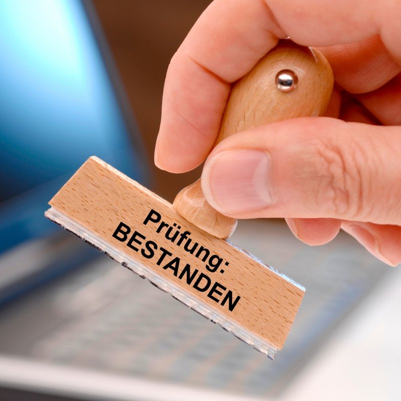 Eine menschliche Hand hält einen Holzstempel mit der Aufschrift „Prüfung: BESTANDEN“. Der Fokus liegt auf dem Stempel, der in den Vordergrund gehalten wird. Im Hintergrund ist unscharf ein aufgeklappter Laptop zu erkennen.  Stempel mit der Aufschrift „Prüfung: BESTANDEN“ wird von einer Hand über einem Laptop gehalten.