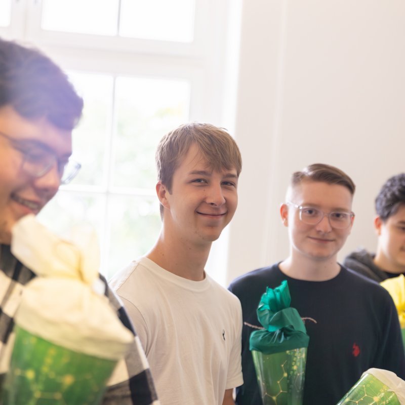Drei junge Menschen stehen nebeneinander in einem Raum und halten grüne Geschenktüten in der Hand. Zwei lächeln in die Kamera, eine Person schaut auf die Geschenktüte. Auf dem Foto stehen drei junge Personen in einem hellen Raum mit großen Fenstern. Alle halten grüne Geschenktüten in Form von Zuckertüten in den Händen. Die linke Person trägt ein weißes T-Shirt und lächelt in Richtung Kamera. Die Person in der Mitte trägt ein dunkles Oberteil und blickt ebenfalls freundlich in die Kamera. Die rechte Person trägt einen dunkelgrauen Kapuzenpullover und betrachtet konzentriert die Zuckertüte in ihren Händen. Im Hintergrund sind Kartons zu sehen, die auf die Situation eines Büro- oder Schulungsraums hindeuten. Die Stimmung wirkt freundlich und entspannt.