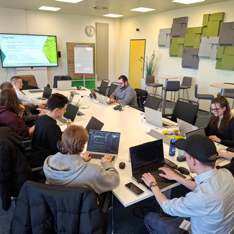 IT-Lehrgang zur objektorientierten Programmierung: Teilnehmende arbeiten gemeinsam an Laptops in einem Schulungsraum In einem hellen Seminarraum sitzen mehrere Auszubildende und Studierende an einem großen Tisch und arbeiten an ihren Laptops. An der Wand ist ein Bildschirm mit Präsentationsfolien zur Programmierung zu sehen. Die Teilnehmenden konzentrieren sich auf praktische Übungen und tauschen sich teilweise miteinander aus. Der Raum ist modern eingerichtet und für Gruppenarbeit ausgelegt.