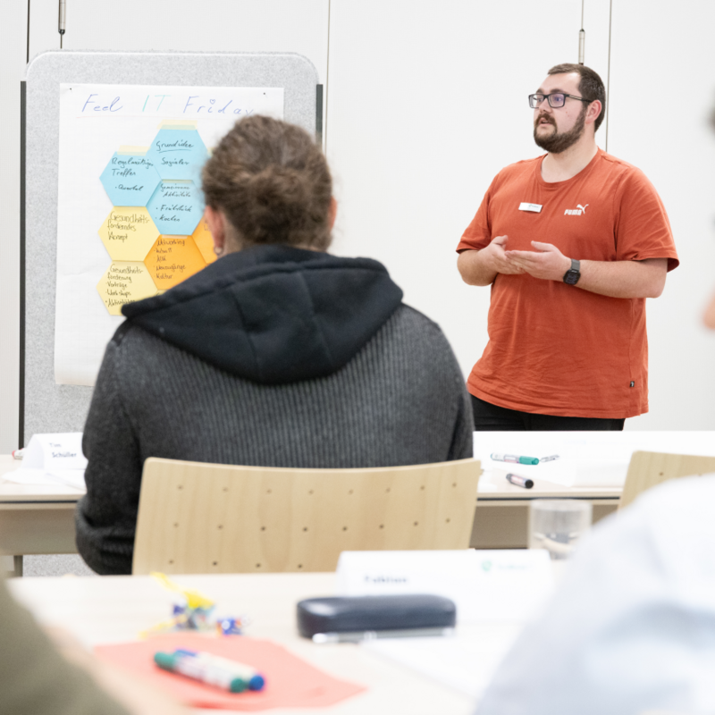 Talent Day 2025 der kubus IT: Austausch, Digitalisierung und Innovation im Fokus Ein junger Auszubildender in rotem Shirt präsentiert beim Talent Day 2025 der kubus IT im Bildungszentrum Hersbruck eine Gruppenarbeit, während Teilnehmende aufmerksam zuhören. Auf einem Flipchart stehen Themen wie Digitalisierung und Arbeitsumfeld.