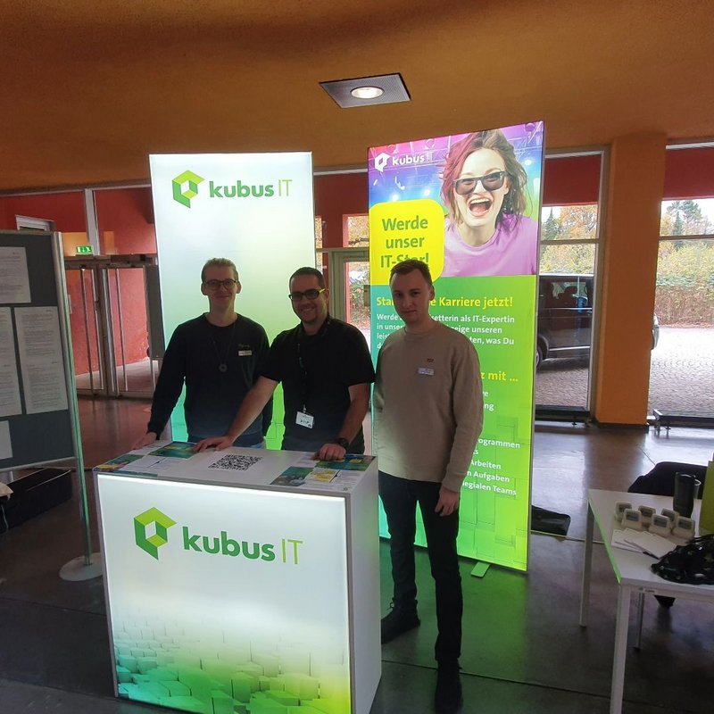 3 Mitarbeiter stehen am Messestand und schauen in die Kamera 3 Mitarbeiter stehen am Messestand und schauen in die Kamera