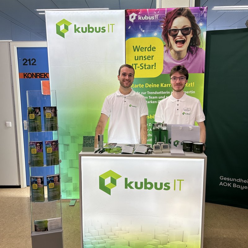 Zwei Personen stehen an einem Messestand der kubus IT. Auf dem beleuchteten Tresen und den Bannern sind das kubus IT Logo und Werbematerialien zu sehen. Das Bild zeigt einen Messestand der kubus IT auf einer Ausbildungsmesse. Zwei Personen stehen hinter einem beleuchteten weißen Tresen mit dem kubus IT Logo in Grün. Beide tragen weiße Poloshirts mit dem Firmenlogo. Rechts auf dem Tresen liegen Werbematerialien wie Flyer, Kugelschreiber, Tassen und Trinkflaschen. Hinter ihnen befinden sich zwei große Banner: eines mit dem kubus IT Logo, das andere mit einem auffälligen Motiv einer jungen Person und dem Slogan „Werde unser IT-Star! Starte Deine Karriere jetzt!“. Links im Bild steht ein Prospektständer mit Flyern.