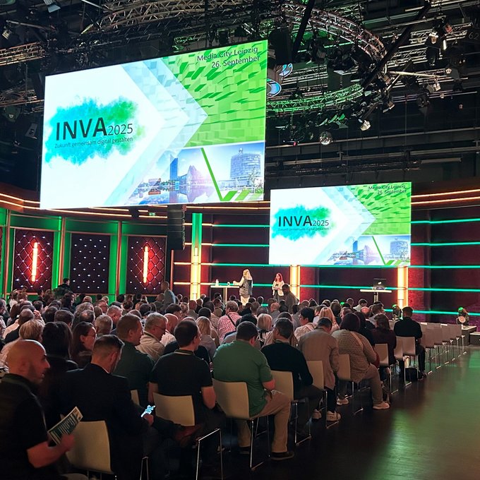 INVA 2025 in Leipzig: Zukunft gemeinsam digital gestalten Großer Veranstaltungssaal mit Bühne und zwei großen Bildschirmen, auf denen „INVA 2025 – Zukunft gemeinsam digital gestalten“ angezeigt wird. Viele Menschen sitzen im Publikum und blicken nach vorne. Die Bühne ist in grün und orange beleuchtet, zwei Moderatorinnen stehen vor der Leinwand.