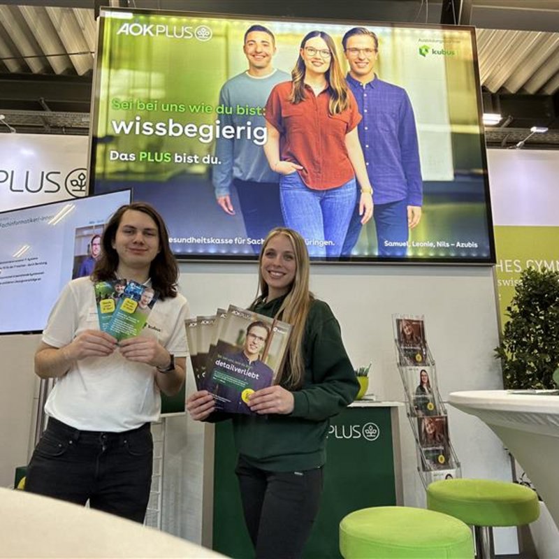 Zwei Ausbilder der kubus IT am Messestand der AOK PLUS auf der Karrierestart Dresden. Ein Mann und eine Frau stehen lächelnd an einem Messestand der AOK PLUS und halten Informationsbroschüren in die Kamera. Im Hintergrund ist ein großer Monitor mit der Aufschrift „Sei bei uns wie du bist: wissbegierig. Das PLUS bist du.“ zu sehen, auf dem drei junge Menschen abgebildet sind. Der Stand ist modern gestaltet, mit grünen Hockern, Informationsmaterialien und dem Logo der AOK PLUS.