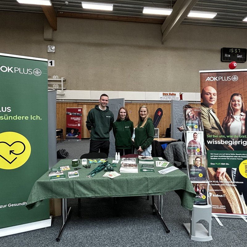 Messestand der AOK PLUS auf einer Berufsmesse mit Informationsmaterialien und mehreren Mitarbeitenden im Gespräch Zu sehen ist ein Messestand der AOK PLUS in einer Halle während einer Berufs- und Studienmesse. Mehrere Mitarbeitende stehen hinter einem Tisch mit Informationsmaterialien und Giveaways und sprechen mit Besucherinnen und Besuchern. Im Hintergrund sind Banner und Roll-ups mit Motiven aus dem Gesundheitswesen sowie weitere Messestände anderer Aussteller zu erkennen. Die Szene vermittelt eine offene, beratende Atmosphäre und zeigt den direkten Austausch mit interessierten Schülerinnen und Schülern.